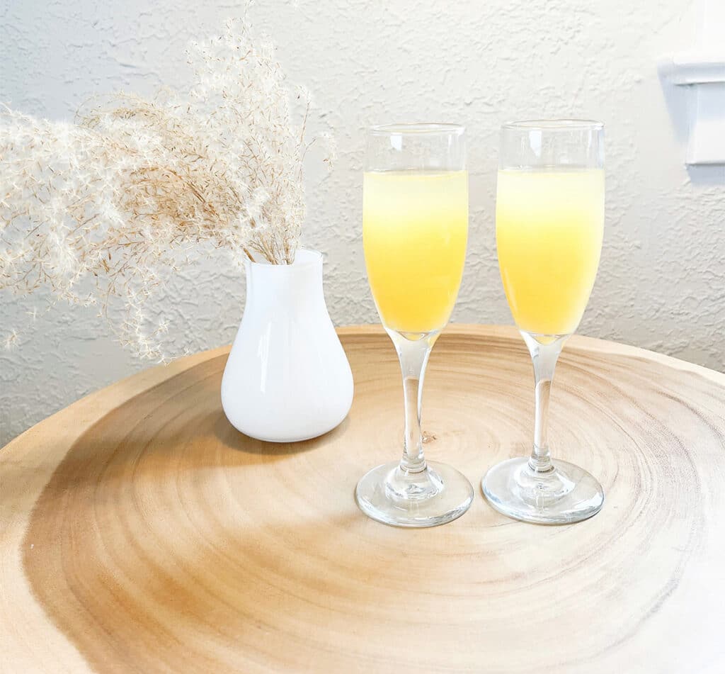 Two Mimosa glasses sitting on a round log table next to a vase of plants.